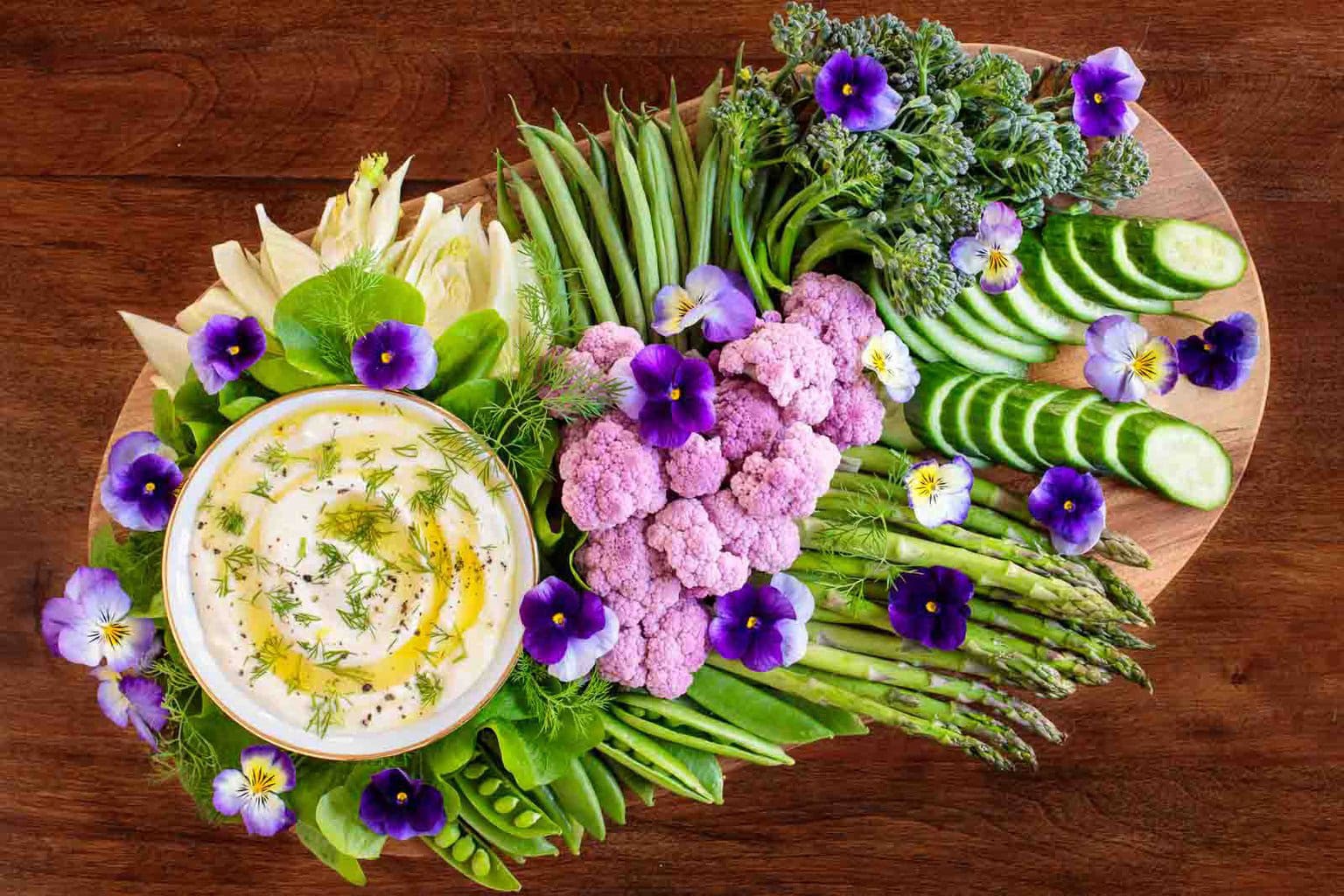 a platter of colorful vegetables and a bowl of Mediterranean Lemon Feta Dip highlighted with purple pansies throughout