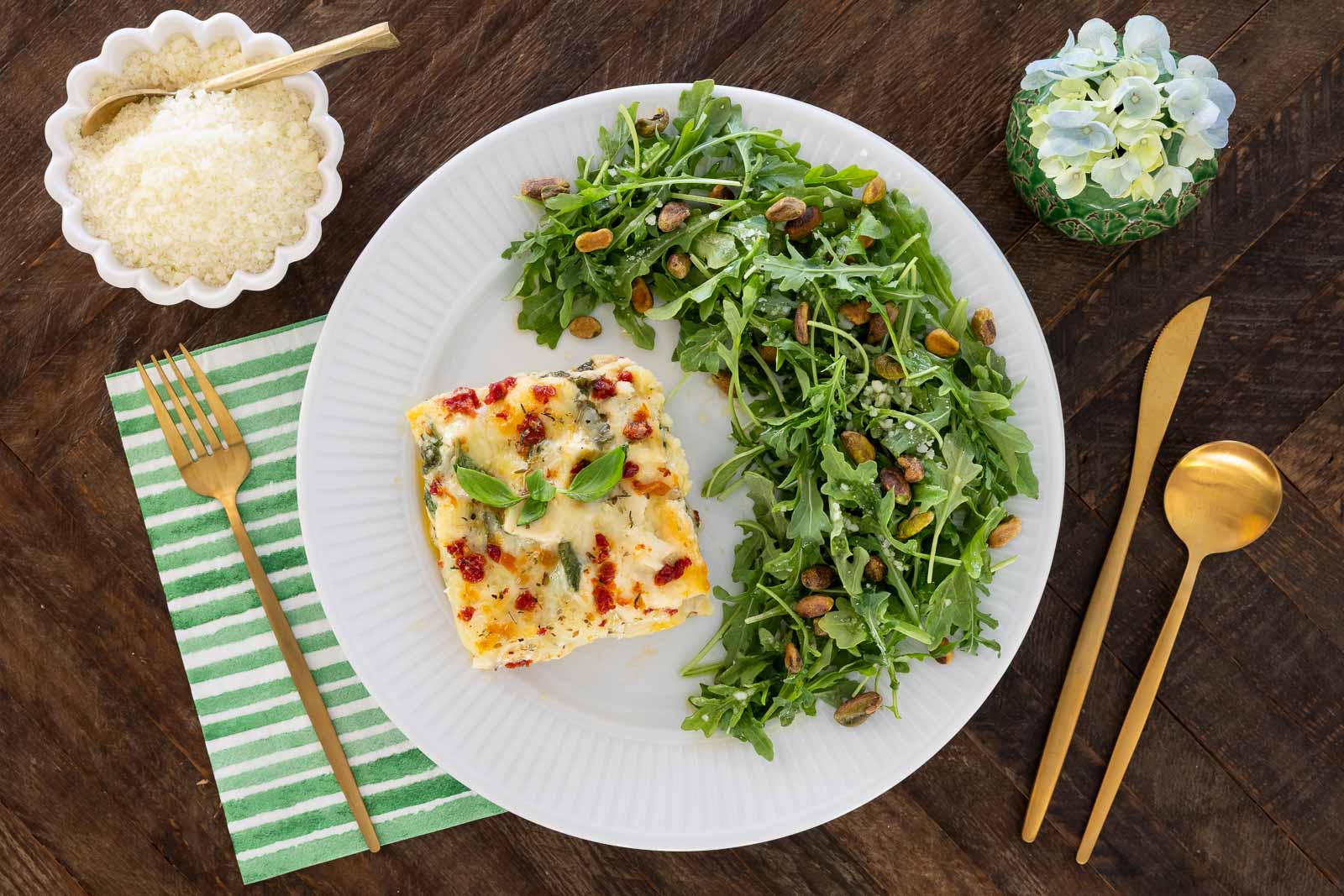 Plated serving of chicken and spinach ravioli lasagna with fresh arugula salad, Parmesan cheese, and gold flatware on a wooden table.