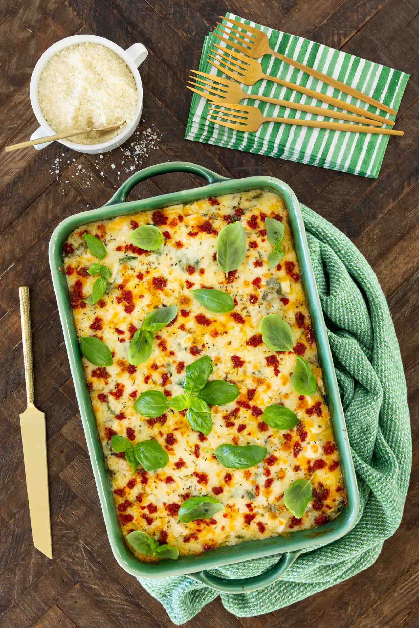 Vertical overhead view of chicken and spinach ravioli lasagna in a green casserole dish topped with melted cheese, sun-dried tomatoes, and fresh basil leaves with Parmesan cheese and napkins nearby.