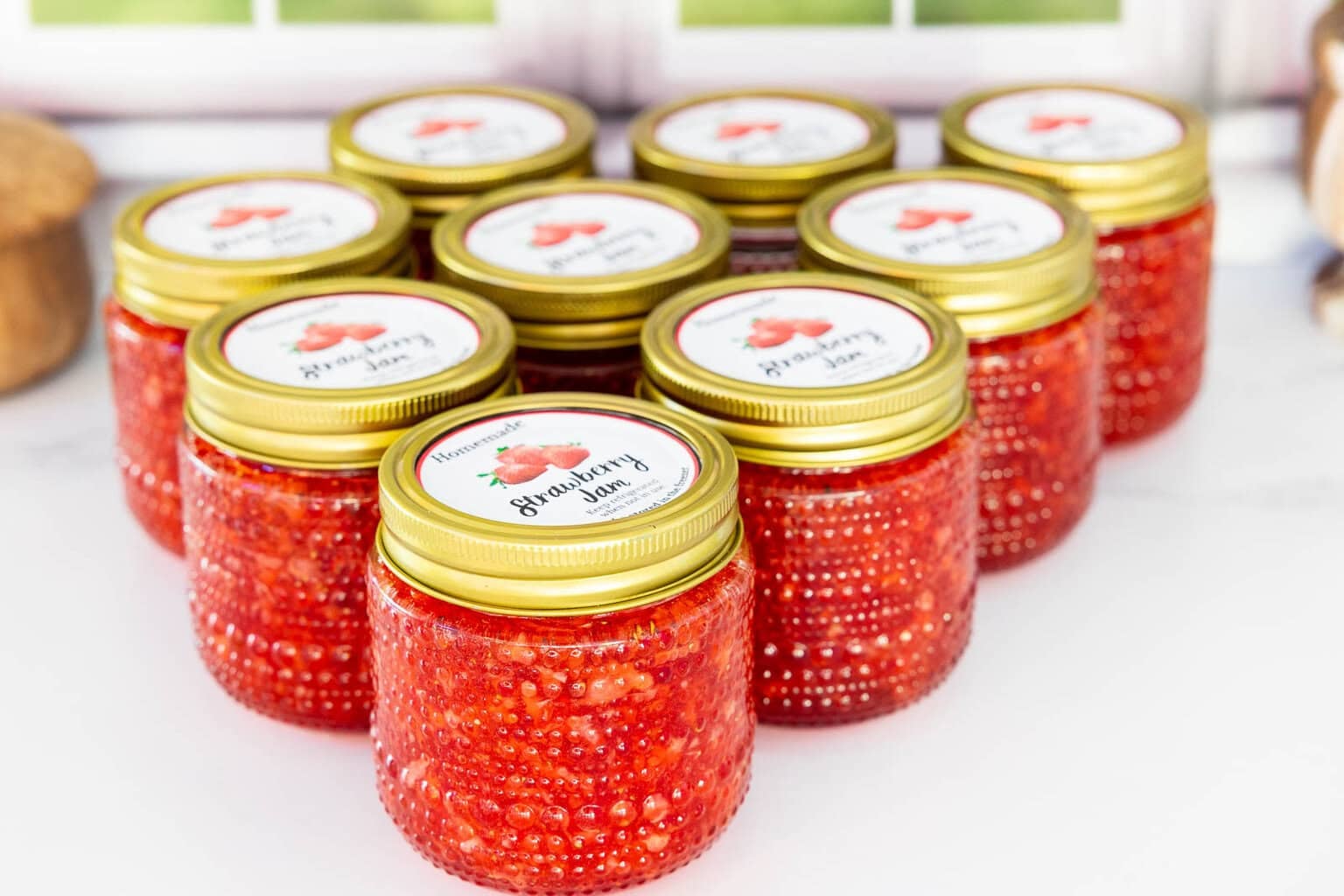 Several small glass jars filled with homemade strawberry freezer jam, topped with gold lids and strawberry jam labels, arranged on a white surface.