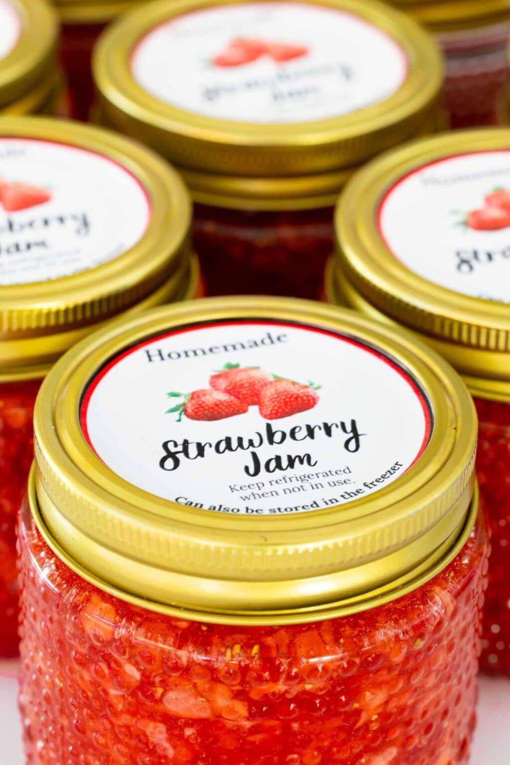 Close-up of small jars filled with homemade strawberry freezer jam topped with gold lids and decorative strawberry jam labels.