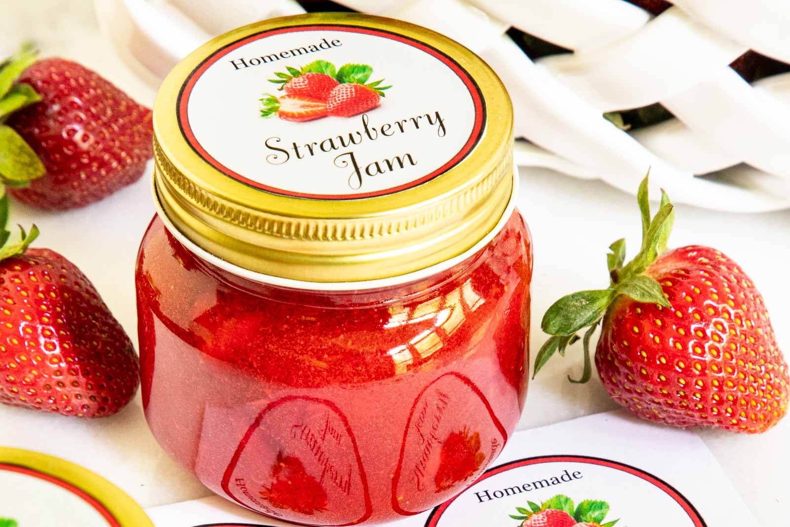 Glass jar of homemade strawberry freezer jam with a gold lid and decorative label, surrounded by fresh strawberries and printable jam labels.