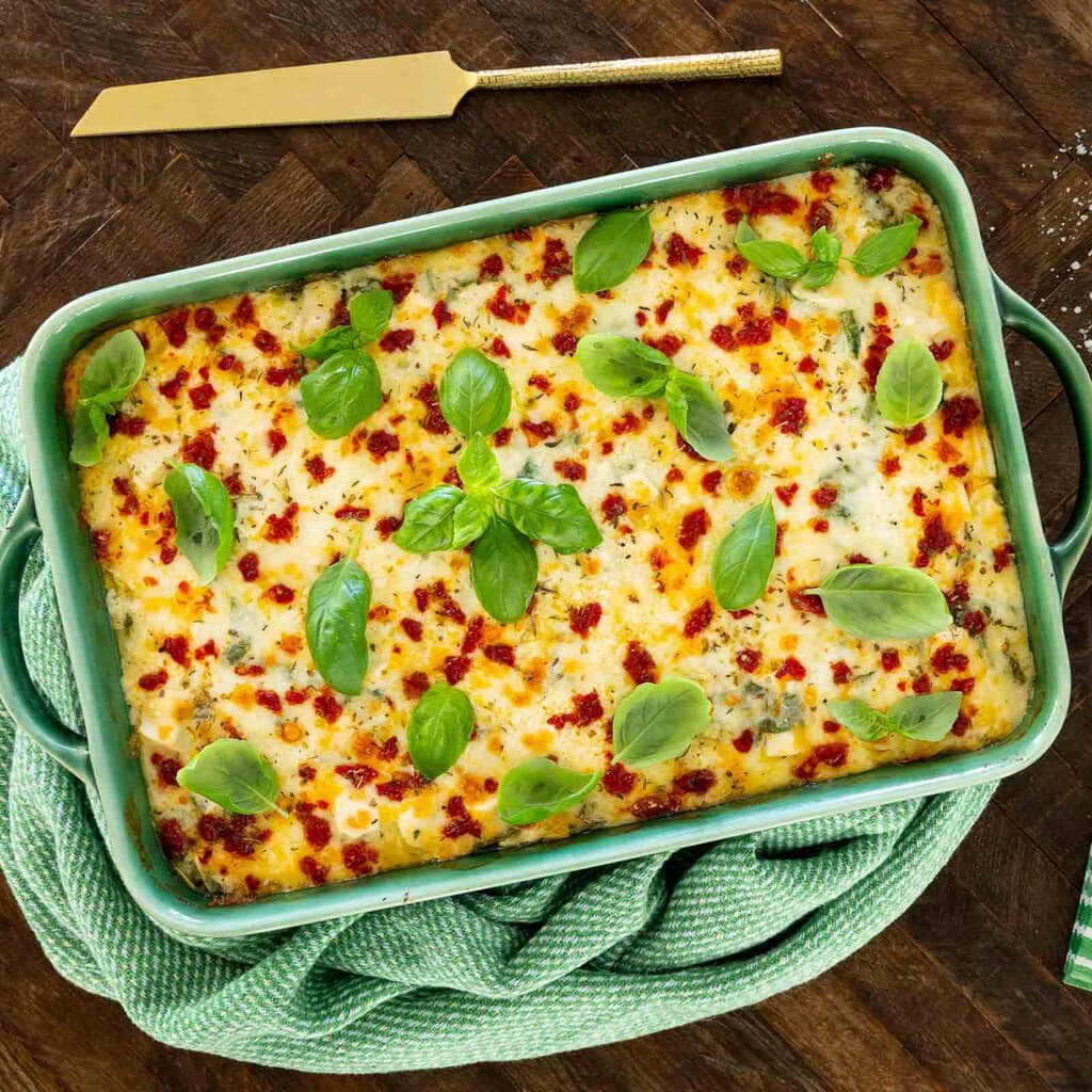 Overhead view of chicken and spinach ravioli lasagna in a green baking dish topped with melted cheese, sun-dried tomatoes, and fresh basil on a wooden table.