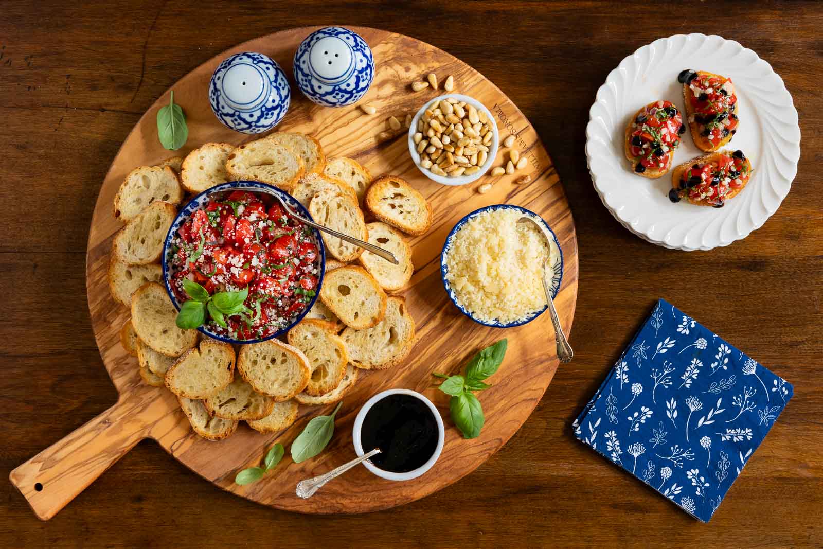 Overhead view of Italian bruschetta with cherry tomato topping, toasted bread slices, Parmesan cheese, balsamic glaze, and pine nuts served on a wooden board.