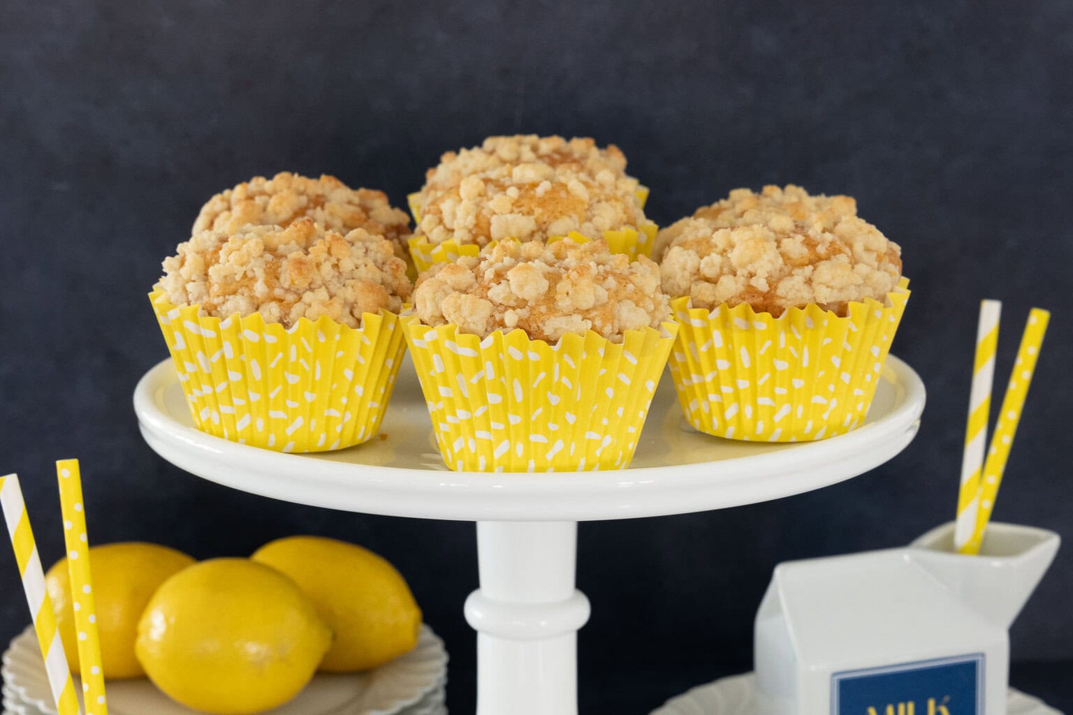 Lemon crumble muffins with buttery streusel topping in yellow liners on a cake stand with fresh lemons