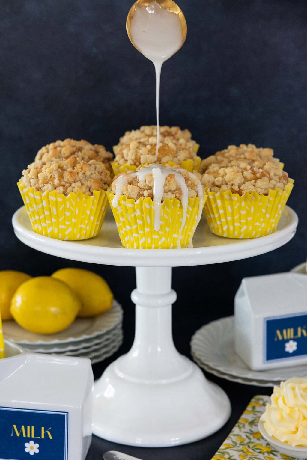 Lemon glaze being drizzled over lemon crumble muffins with streusel topping on a cake stand