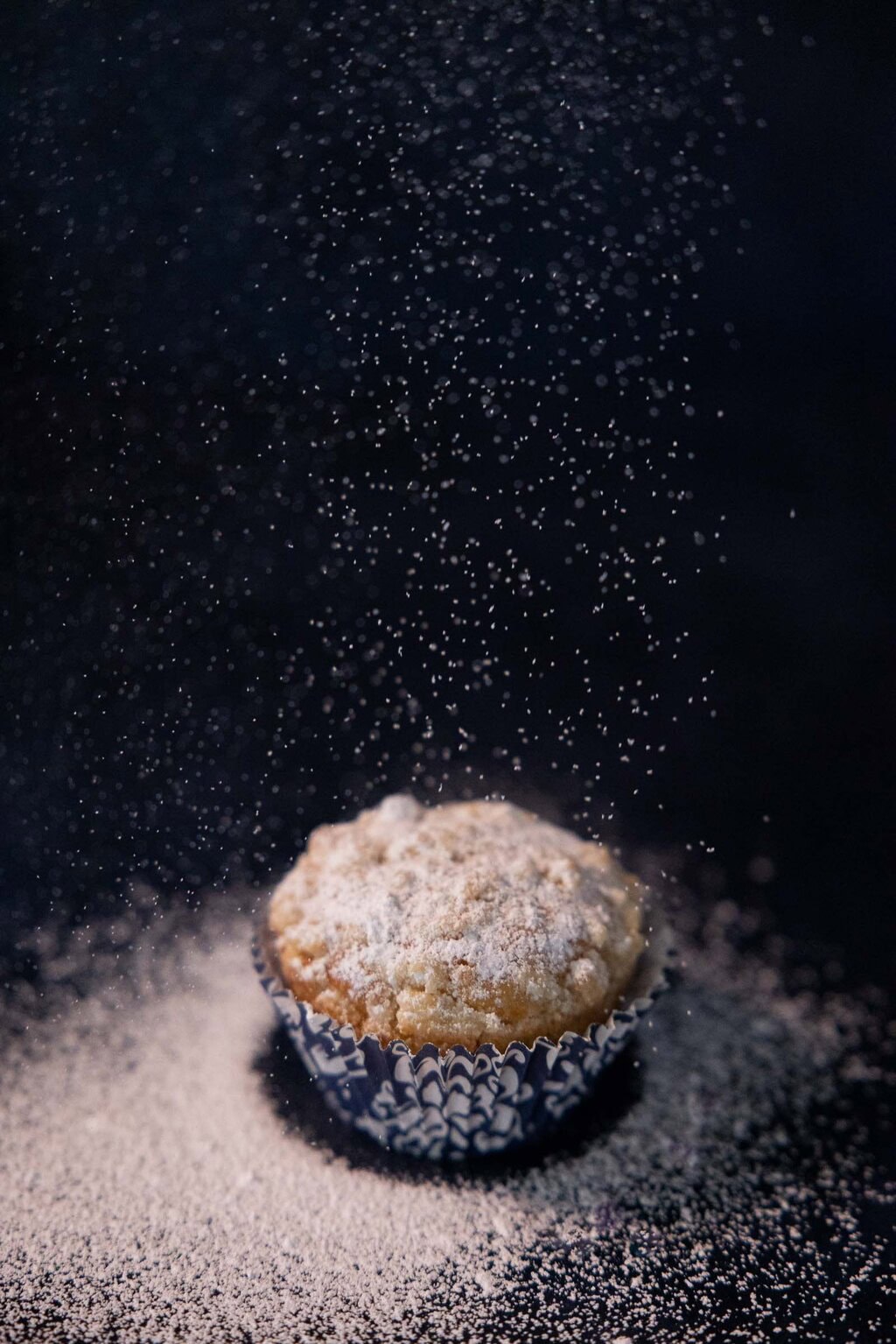 Single lemon crumble muffin dusted with powdered sugar on a dark surface with falling sugar