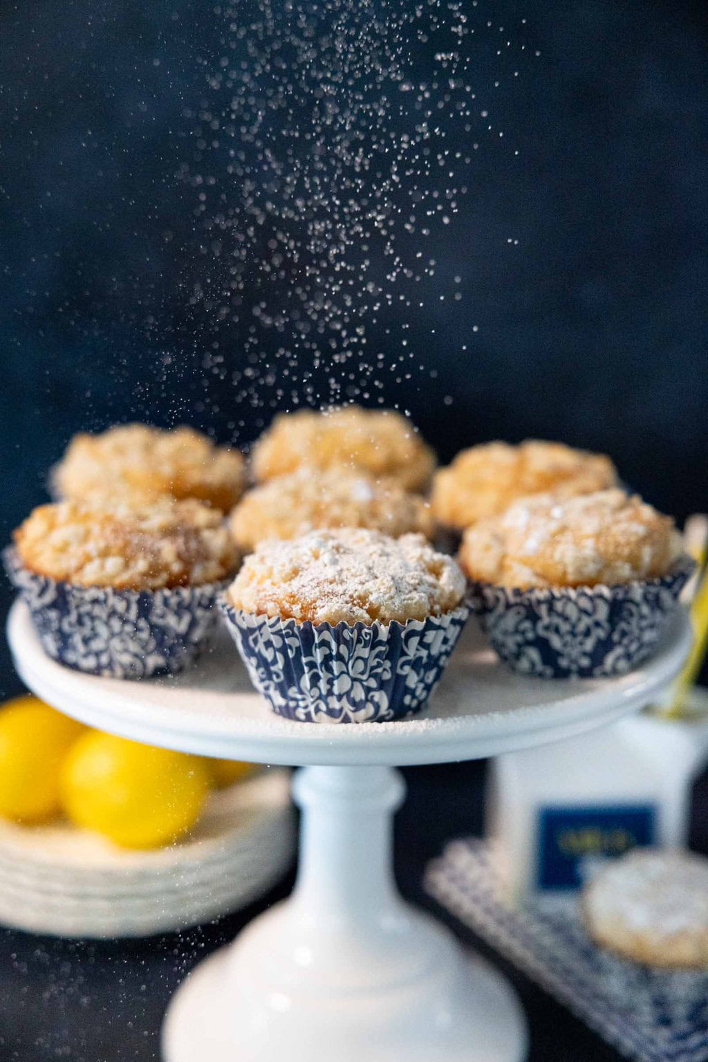 Powdered sugar being dusted over lemon crumble muffins with streusel topping on a cake stand