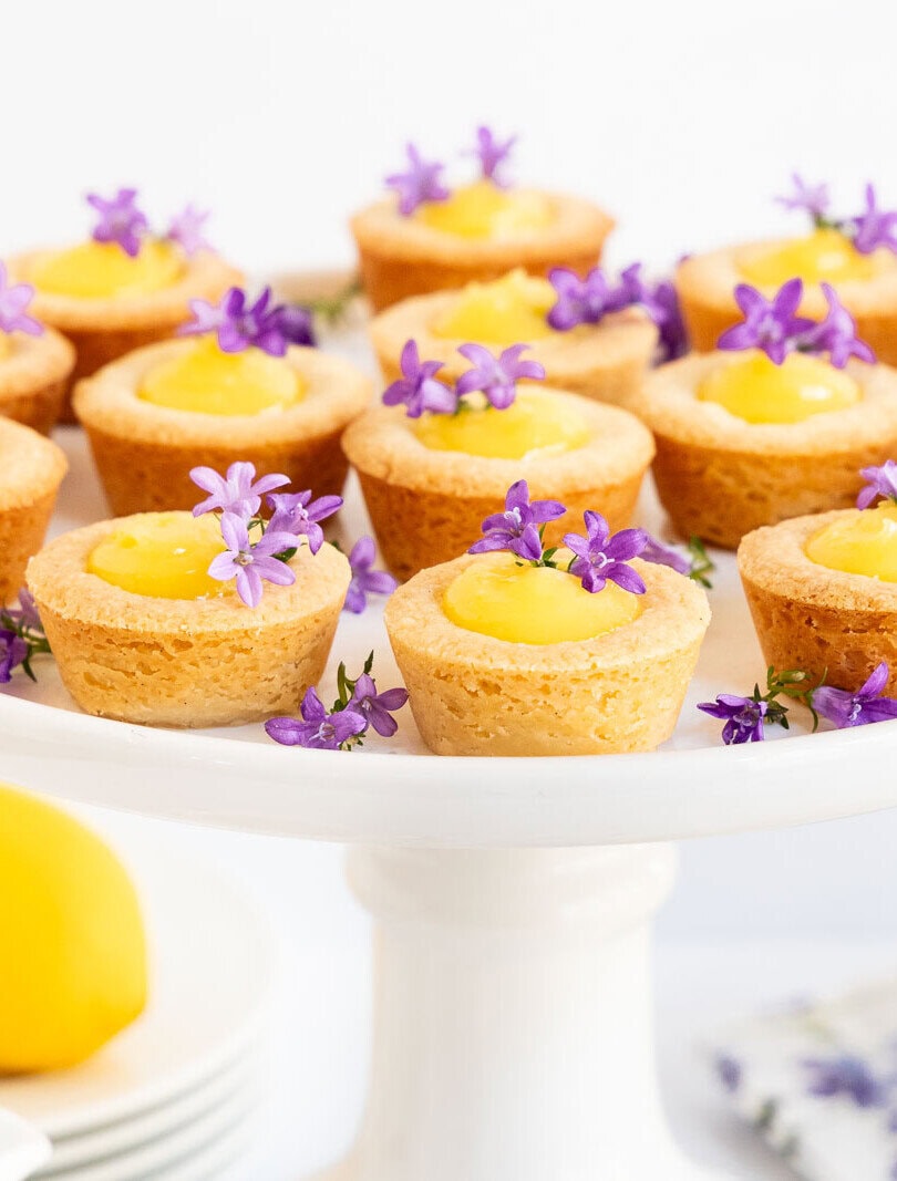 Mini lemon curd tartlets with shortbread crusts on a white cake stand, topped with purple flowers and served with fresh lemons.
