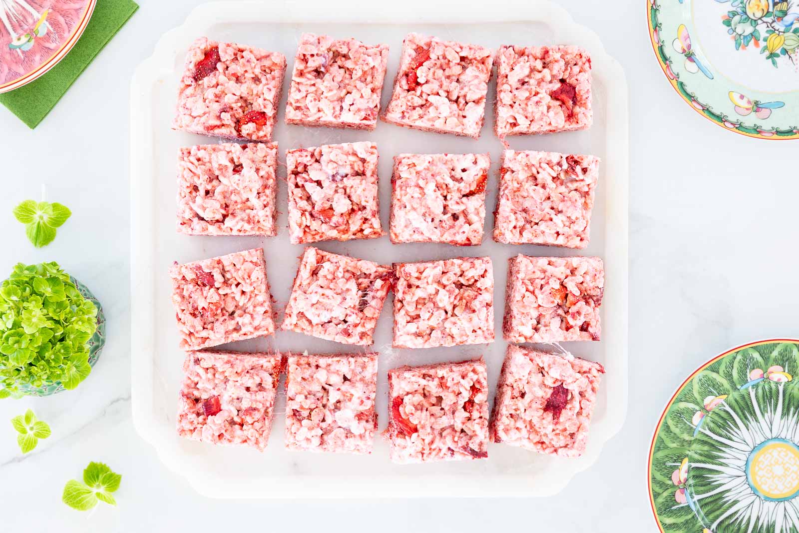 Overhead view of strawberry rice krispie treats cut into squares on a tray with decorative plates and fresh green accents