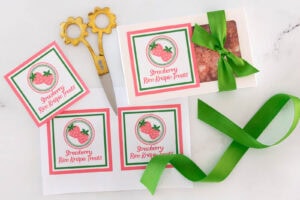 Printable strawberry Rice Krispie treat labels with gift box tied with green ribbon and kitchen scissors on marble surface