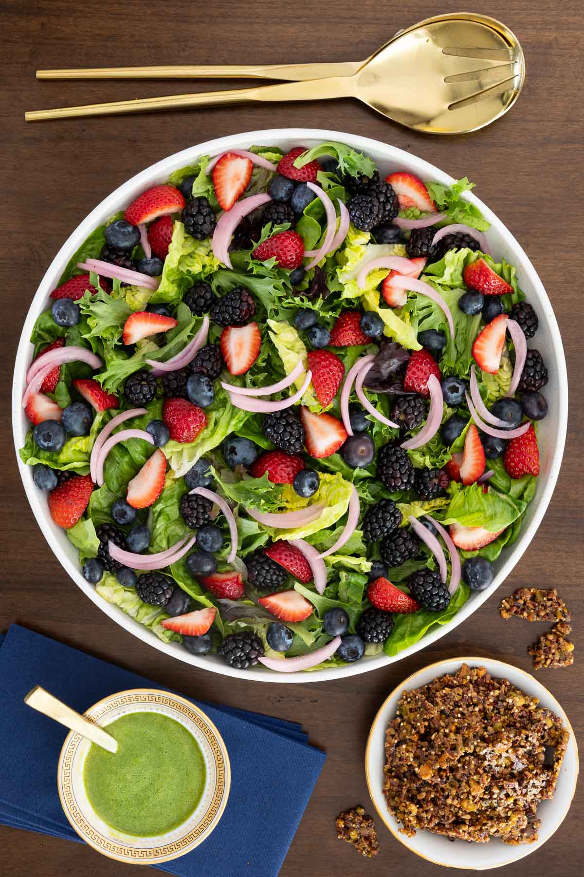 Overhead view of a Summer Berry Green Salad with strawberries, blueberries, blackberries, pickled red onions, Honey Lemon Mint Dressing and crunchy Maple Pistachio Brittle on a wooden table.