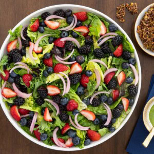 Overhead view of a Summer Berry Green Salad with strawberries, blueberries, blackberries, pickled red onions, Honey Lemon Mint Dressing and crunchy Maple Pistachio Brittle on a wooden table.