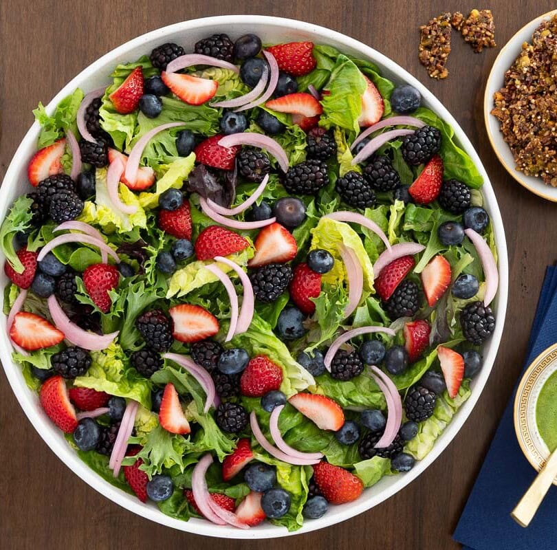 Overhead view of a Summer Berry Green Salad with strawberries, blueberries, blackberries, pickled red onions, Honey Lemon Mint Dressing and crunchy Maple Pistachio Brittle on a wooden table.