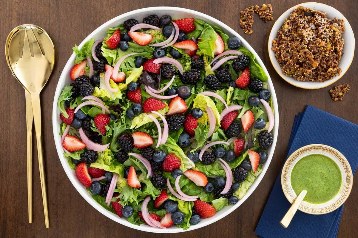 Overhead view of a Summer Berry Green Salad with strawberries, blueberries, blackberries, pickled red onions, Honey Lemon Mint Dressing and crunchy Maple Pistachio Brittle on a wooden table.