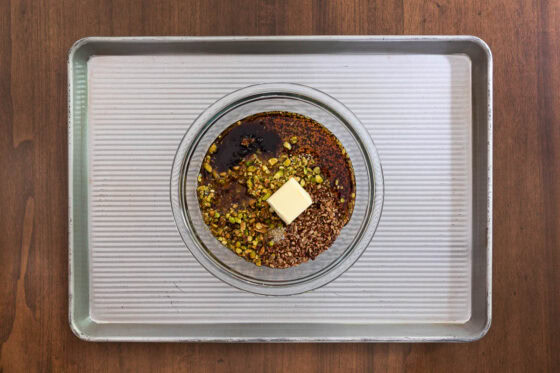 Overhead view of Maple Pistachio Brittle ingredients combined in a glass bowl with maple syrup, pistachios, pecans, quinoa, butter, brown sugar and salt before mixing.