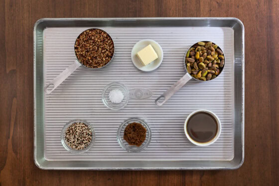 Overhead view of ingredients for Maple Pistachio Brittle including pistachios, chopped pecans, quinoa, maple syrup, brown sugar, butter and salt on a sheet pan.