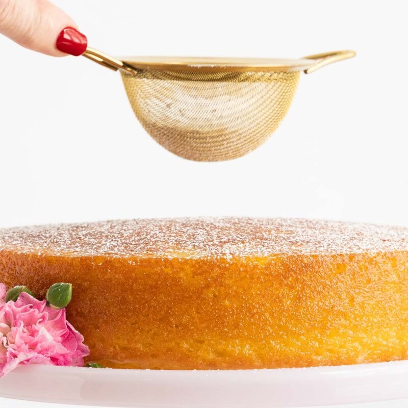Hand dusting powdered sugar over a simple French Butter Cake decorated with pink flowers on a white cake stand.