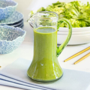 Glass cruet filled with vibrant green Honey Lemon Mint Salad Dressing on a table with blue dishes and gold flatware in the background.
