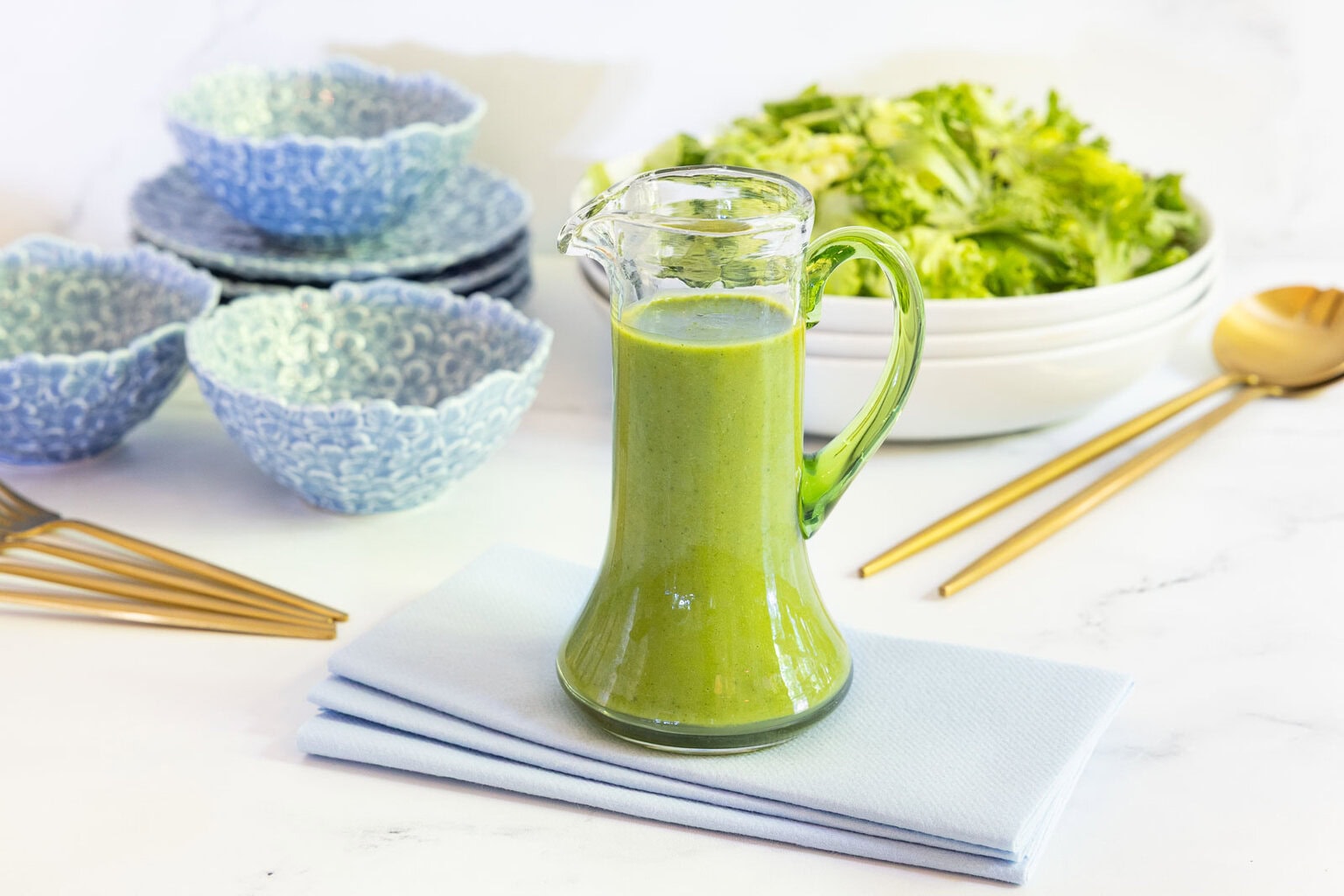 Glass cruet filled with bright green Honey Lemon Mint Salad Dressing on a folded blue napkin with salad bowls and gold flatware in the background.