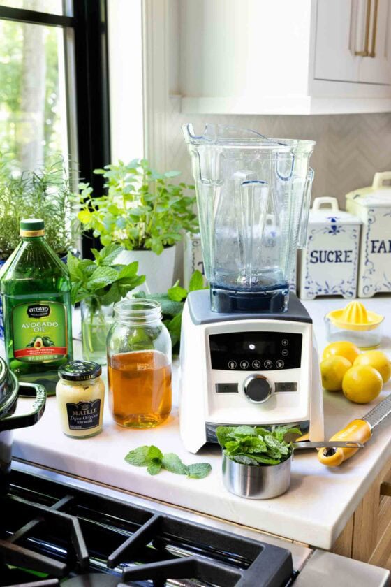 Ingredients for Honey Lemon Mint Salad Dressing arranged beside a blender, including fresh mint, lemons, honey, Dijon mustard and avocado oil on a kitchen counter.