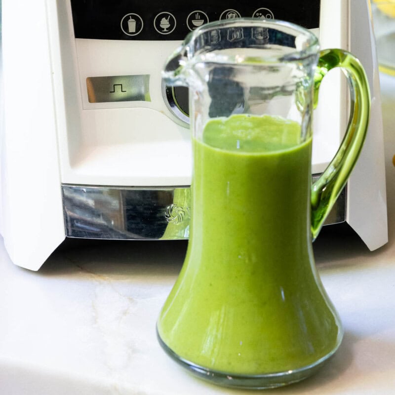 Glass cruet filled with bright green Honey Lemon Mint Salad Dressing beside lemons and a blender on a kitchen counter.
