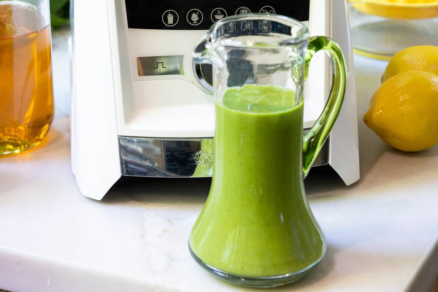 Glass cruet filled with bright green Honey Lemon Mint Salad Dressing beside lemons and a blender on a kitchen counter.