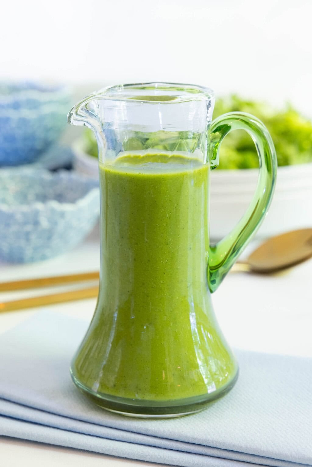 Glass cruet filled with bright green Honey Lemon Mint Salad Dressing on a folded blue napkin with salad bowls and gold flatware in the background.