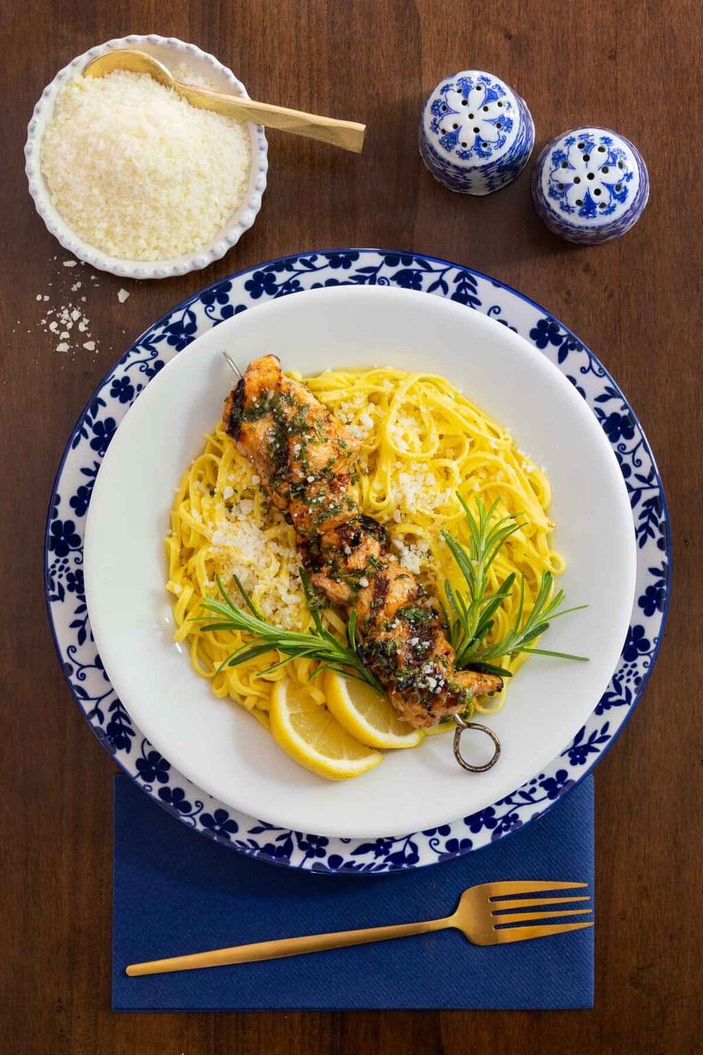Italian chicken skewer served over linguine pasta with grated Parmesan, lemon wedges and fresh rosemary on a blue-and-white Italian-style plate with a navy napkin and gold fork.