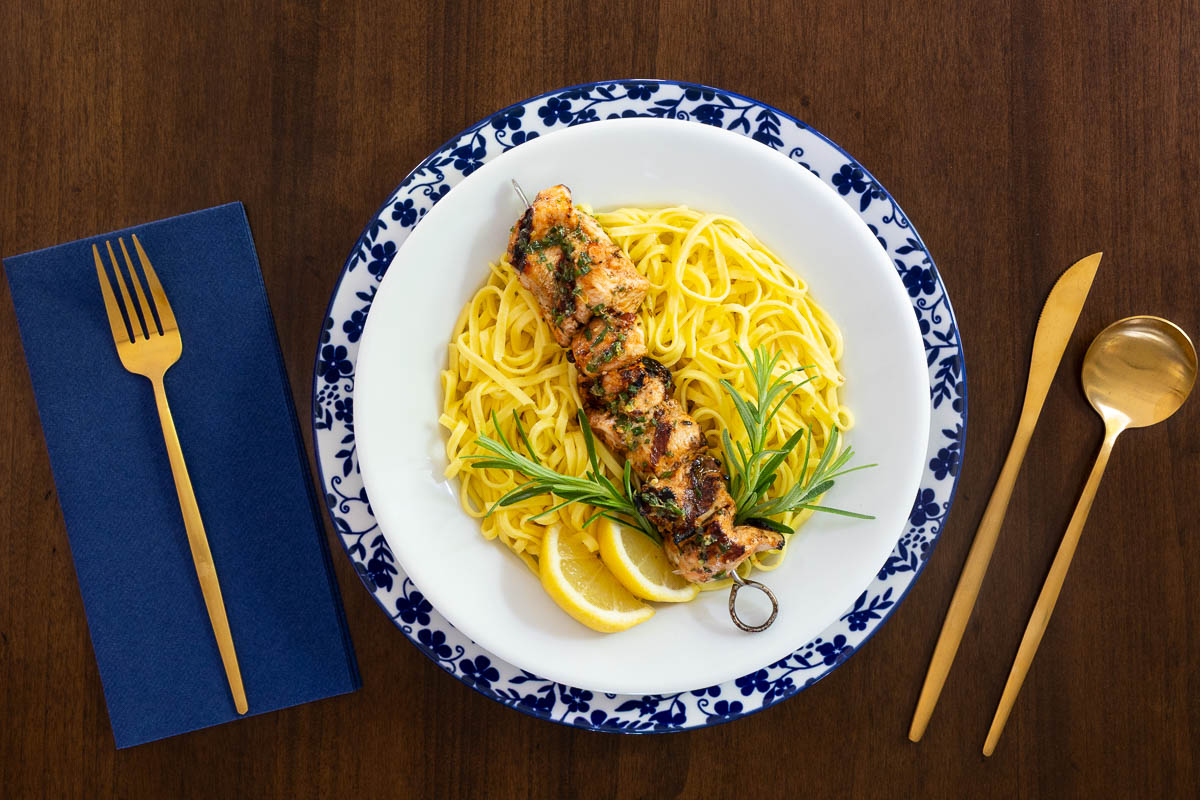 Italian chicken skewer served over fresh linguine pasta with lemon wedges and rosemary on a blue-and-white Italian-style plate, styled with gold flatware and a navy napkin.
