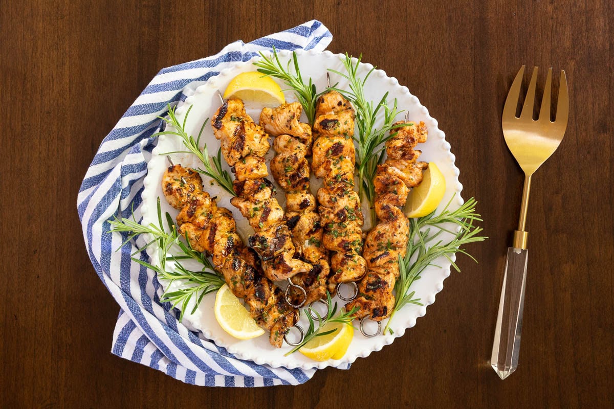 Platter of grilled Italian chicken skewers garnished with fresh rosemary sprigs and lemon wedges on a white serving plate with a blue-striped napkin and gold serving fork.