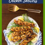 Platter of grilled Italian chicken skewers garnished with fresh rosemary sprigs and lemon wedges on a white serving plate with a blue-striped napkin and gold serving fork.