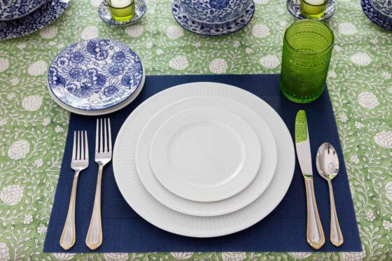 Simple spring place setting with layered white dinnerware, blue placemat, green glass tumbler and blue and white accent plates on a green floral tablecloth.