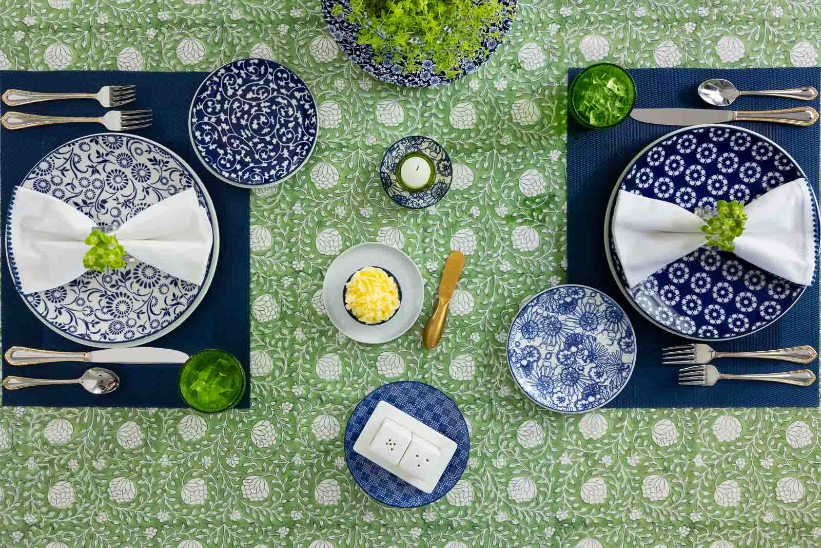 Overhead view of a blue and green spring-summer tablescape with navy placemats, layered blue and white dishes, herb centerpiece and elegant entertaining details.