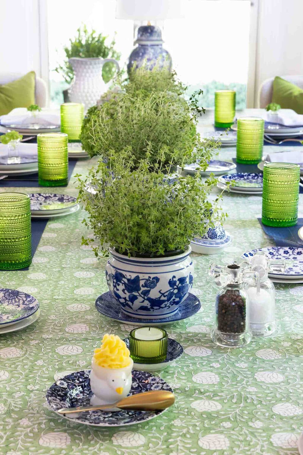 Spring-summer tablescape with potted herb centerpieces in blue and white ginger jars, green glassware, patterned dishes and decorative butter served in a small chicken dish.