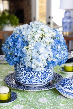 Blue hydrangea centerpiece in a blue and white ceramic bowl on a green patterned tablecloth with blue and white dishes and green candle holders.