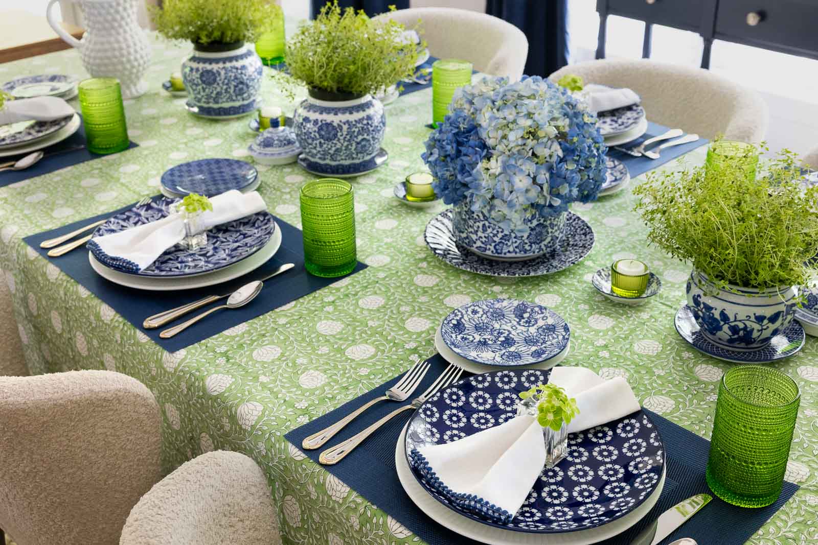 Blue and green spring-summer tablescape with hydrangeas, herb centerpieces, navy placemats, green glassware and layered blue and white dishes.