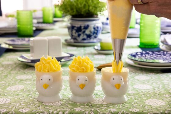 Soft butter being piped into white chick-shaped egg cups on a spring tablescape with blue and white dishes, green glassware and floral accents.