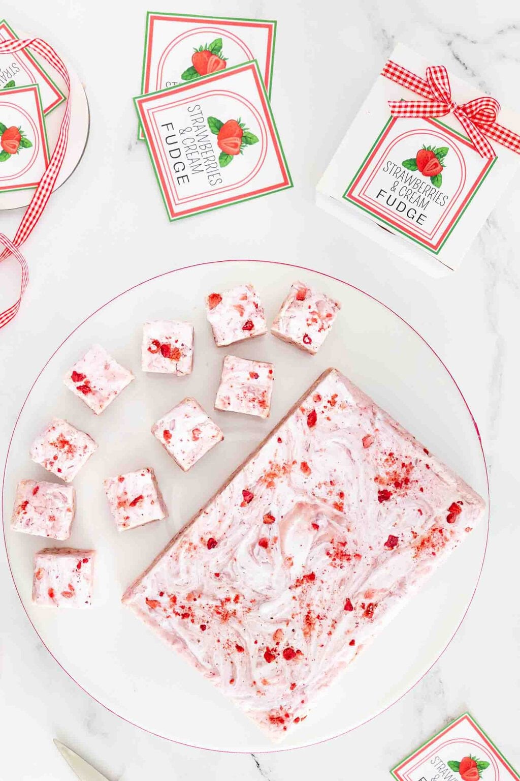 Overhead view of homemade Strawberries & Cream Fudge with swirls of marshmallow creme and crushed strawberries, surrounded by printable gift labels, ribbon and bakery boxes on a white marble background.