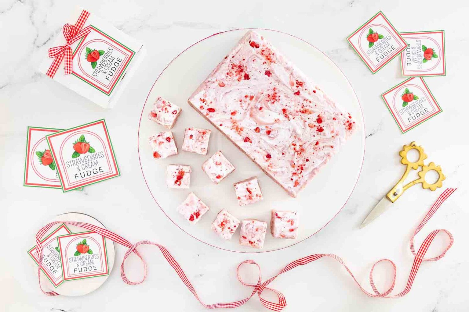Overhead view of homemade Strawberries & Cream Fudge on a white platter with bakery boxes, printable gift labels, ribbon and scissors on a marble background.