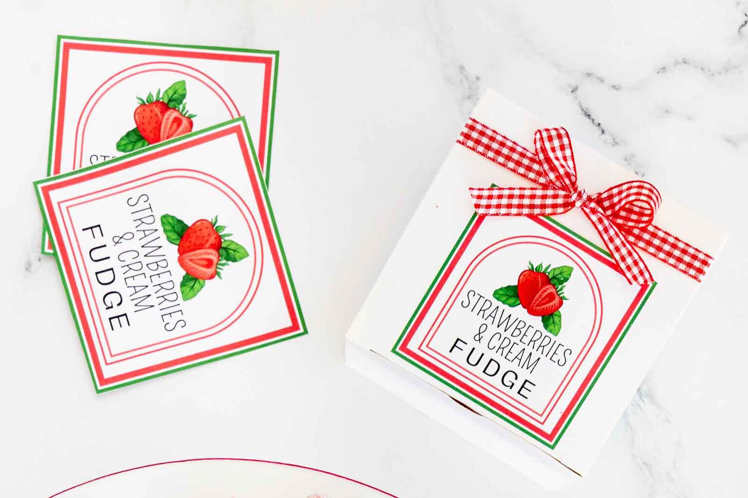 Close-up view of free printable Strawberries & Cream Fudge gift labels attached to a white bakery box with red gingham ribbon on a marble background.