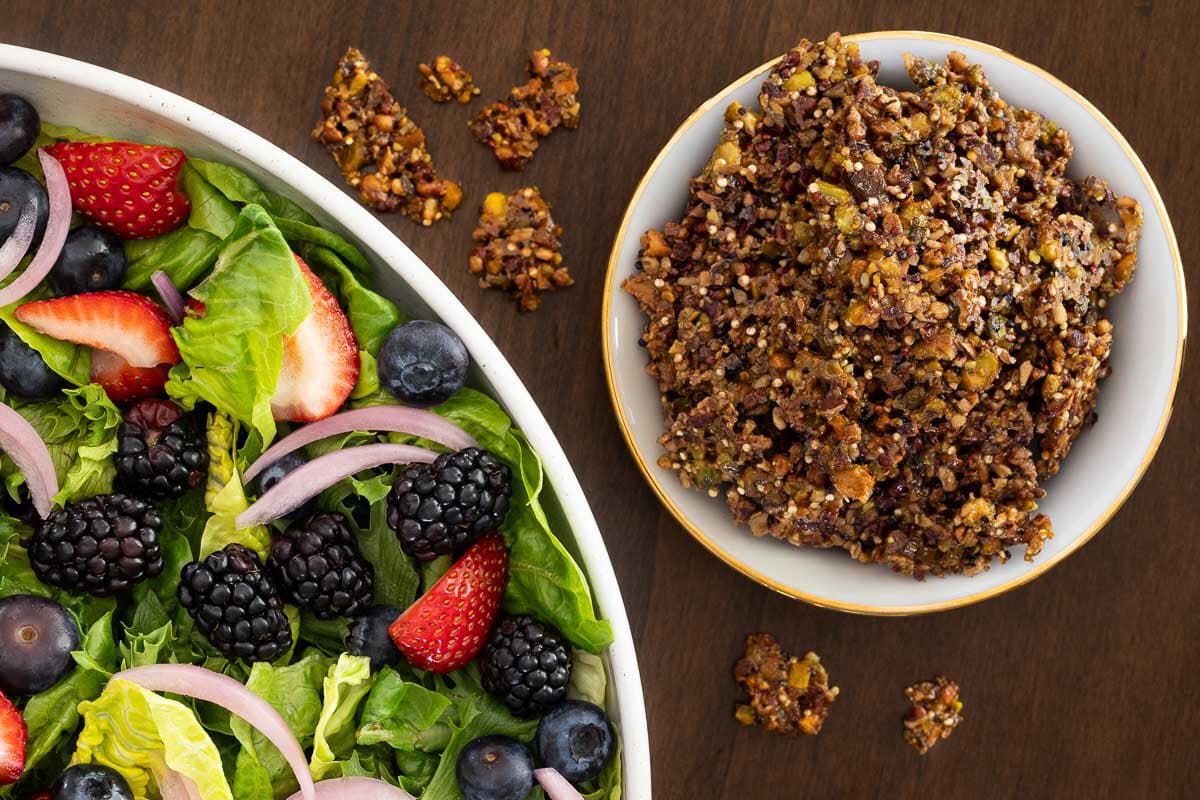 Close-up overhead view of a Summer Berry Green Salad with blackberries, blueberries, strawberries, pickled red onions and a bowl of crunchy Maple Pistachio Brittle.
