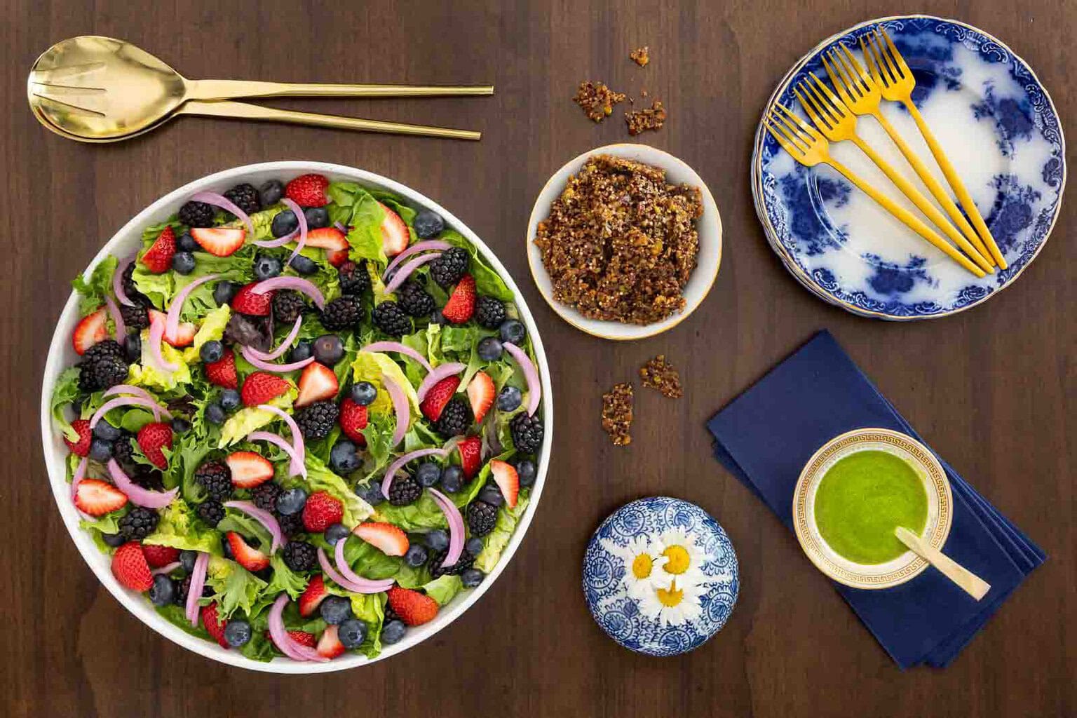Overhead view of a Summer Berry Green Salad with strawberries, blueberries, blackberries and pickled red onions served with maple pistachio brittle and bright green Honey Lemon Mint Dressing.
