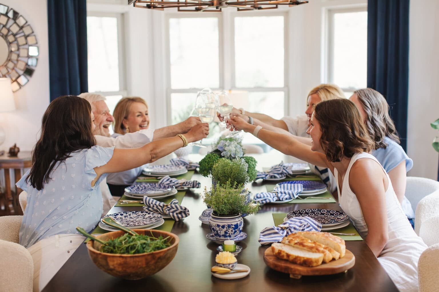 Family and friends gathered around a blue and white summer tablescape raising wine glasses for a toast with fresh flowers, herb centerpieces and elegant dinner party decor.