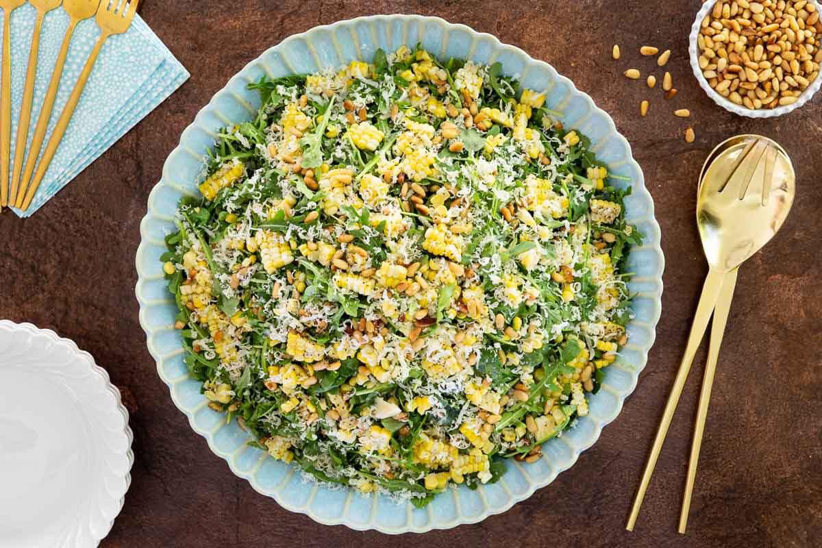 Overhead view of an arugula corn salad with fresh corn, Parmesan cheese, and toasted pine nuts in a blue serving bowl.