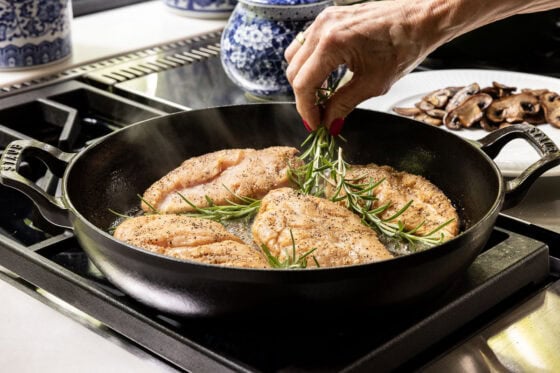 Fresh rosemary sprigs being added to chicken breasts as they sear in a cast iron skillet for One-Pan Balsamic Rosemary Chicken.