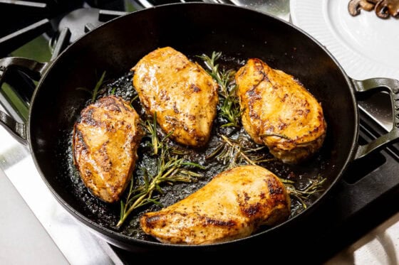 Chicken breasts searing with fresh rosemary in a cast iron skillet for One-Pan Balsamic Rosemary Chicken.