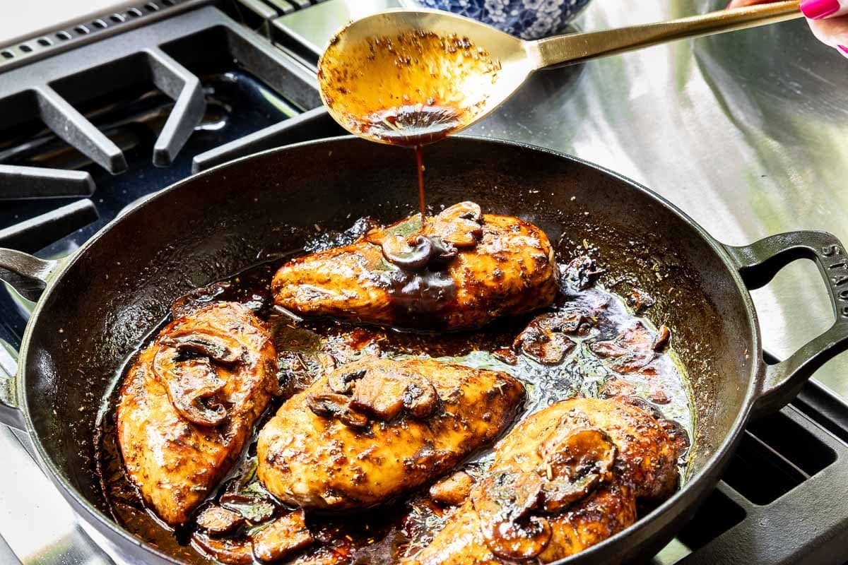 Rich balsamic rosemary pan sauce being spooned over seared chicken breasts and mushrooms in a cast iron skillet.