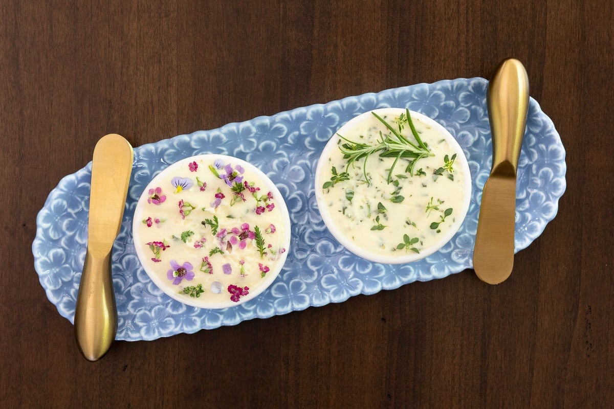 Overhead view of herb butter and flower-topped compound butter on a blue serving tray with gold butter knives.