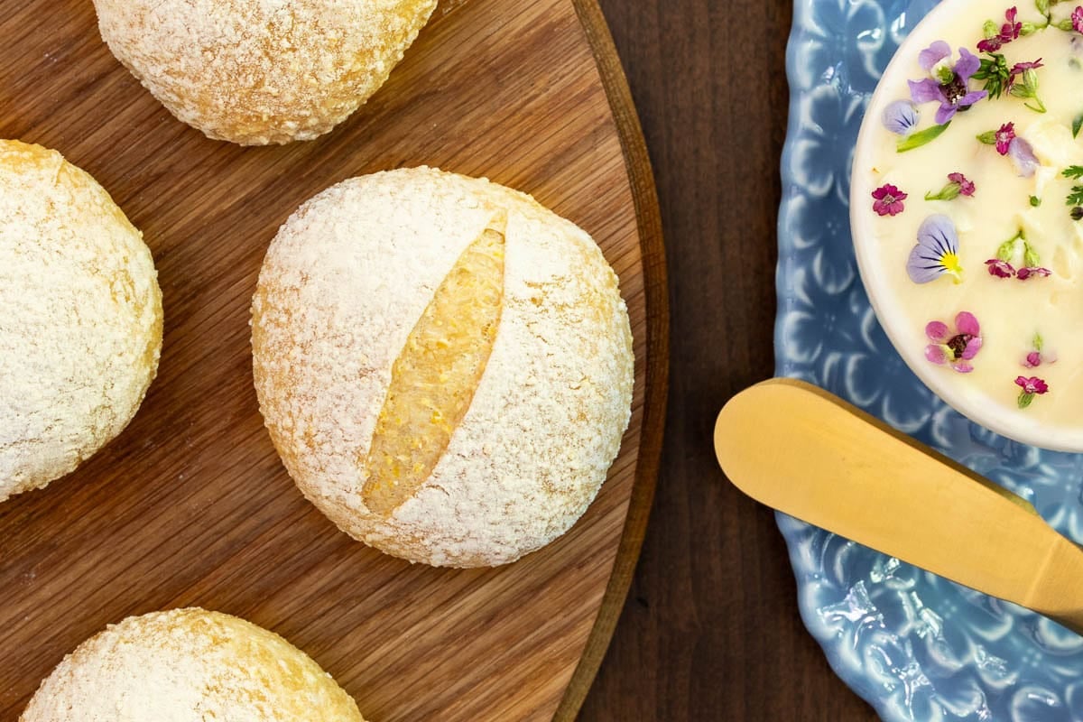 Overhead view of a homemade no-knead Italian-style dinner roll on a wooden serving board with flower-topped compound butter and a gold butter knife.